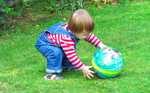 Дете в деним гащеризон играе на тревата с топката